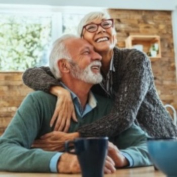 A mature couple smiling and embracing at a wood table in a kitchen.